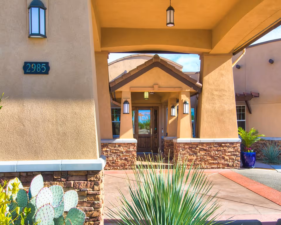 Front entrance of a building with beige stucco walls and stone accents, featuring a covered porch with hanging lantern lights, a wooden door, and desert plants including a cactus and a spiky green plant in the foreground.