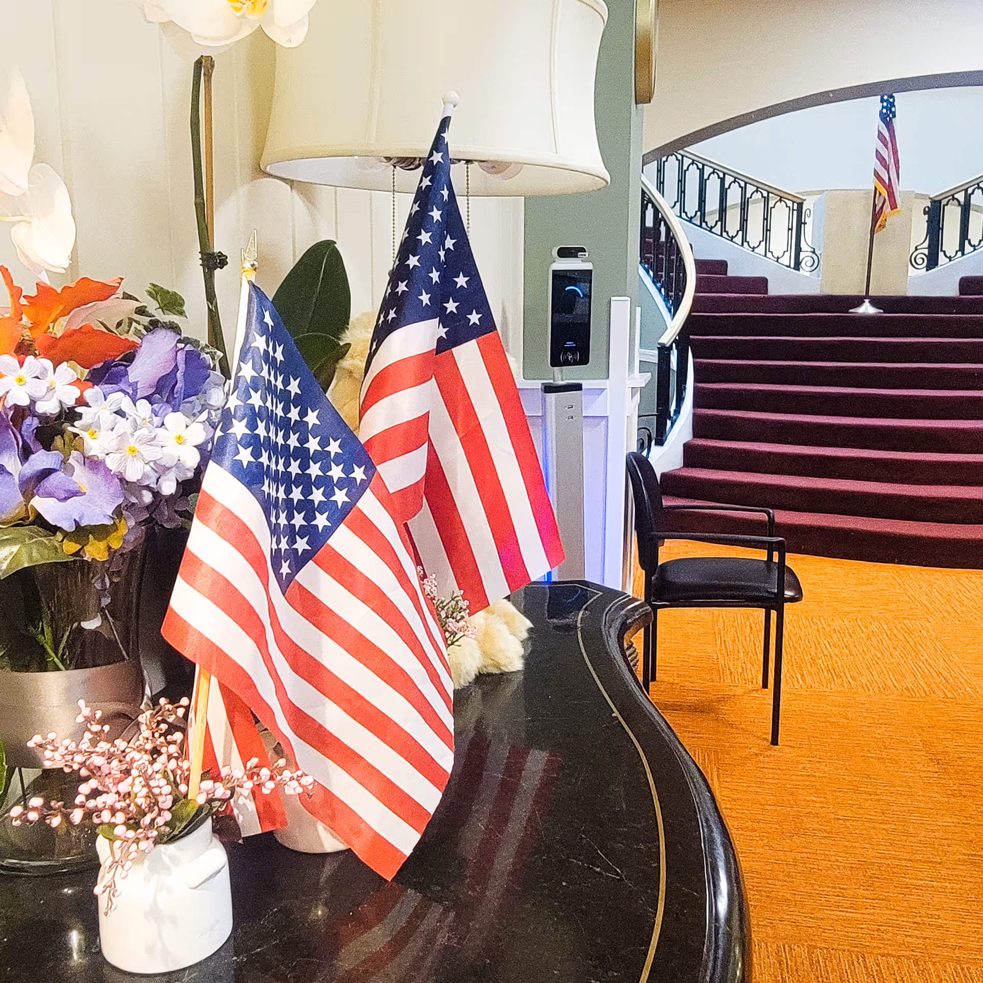 Interior view of a senior living community featuring a black table with two small American flags and flower arrangements. In the background, there is a curved staircase with maroon carpeting and an American flag at the top. A black chair is positioned near the table, and a lamp with a white shade is visible behind the flags.