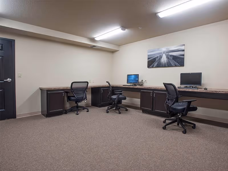 A small office room with beige walls and carpeted floor, featuring a long L-shaped desk with two computer monitors and three black office chairs on wheels. A black door is visible on the left side, and a black and white framed photograph hangs on the wall above the desk.