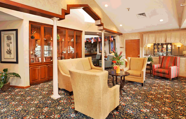A cozy living room area in a senior living facility featuring beige upholstered armchairs and a sofa arranged around a dark wooden coffee table with a flower arrangement. The room has a patterned carpet, wooden display cabinets with glass doors, framed artwork on the walls, and a striped armchair near a window with curtains. The ceiling has recessed lighting and decorative columns.