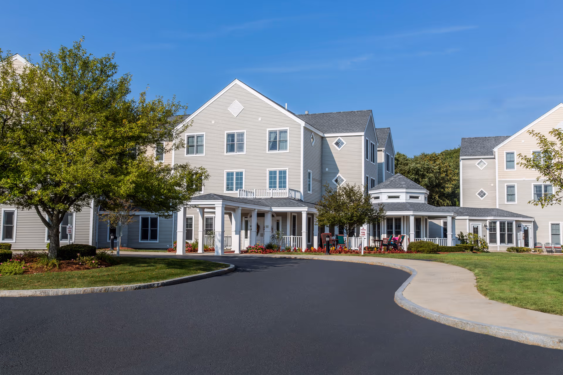 Exterior view of a senior living facility named Benchmark at North Andover, showing a multi-story beige building with white trim, surrounded by green lawns, trees, and a curved driveway under a clear blue sky.