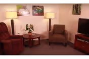 Small living area with two armchairs, a side table with a plant, two floor lamps, a TV on a stand, and framed artwork on the wall.