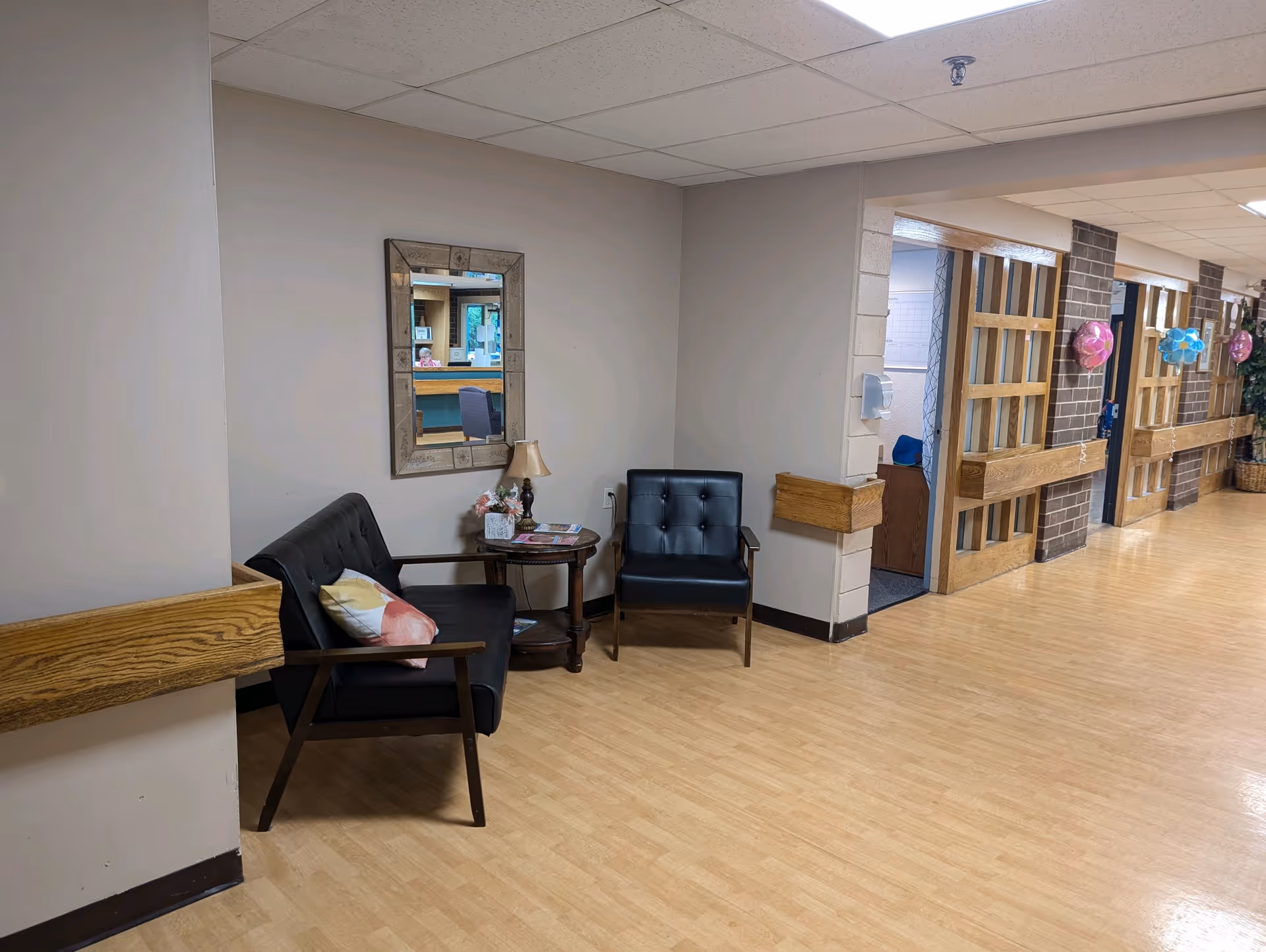 A hallway in a senior living facility with light wood flooring and beige walls. There are two black cushioned chairs with wooden arms, a small round wooden table with a lamp and some magazines, and a decorative mirror on the wall. The hallway has wooden handrails and several doorways with wooden frames and brick accents. Some colorful balloon decorations are visible on the right side of the hallway.