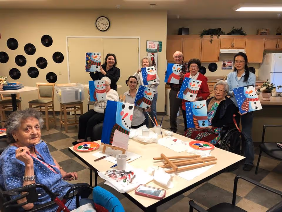 A group of elderly people and caregivers in a communal room holding up paintings of snowmen they created. The room has a kitchen area in the background and tables with painting supplies in the foreground.