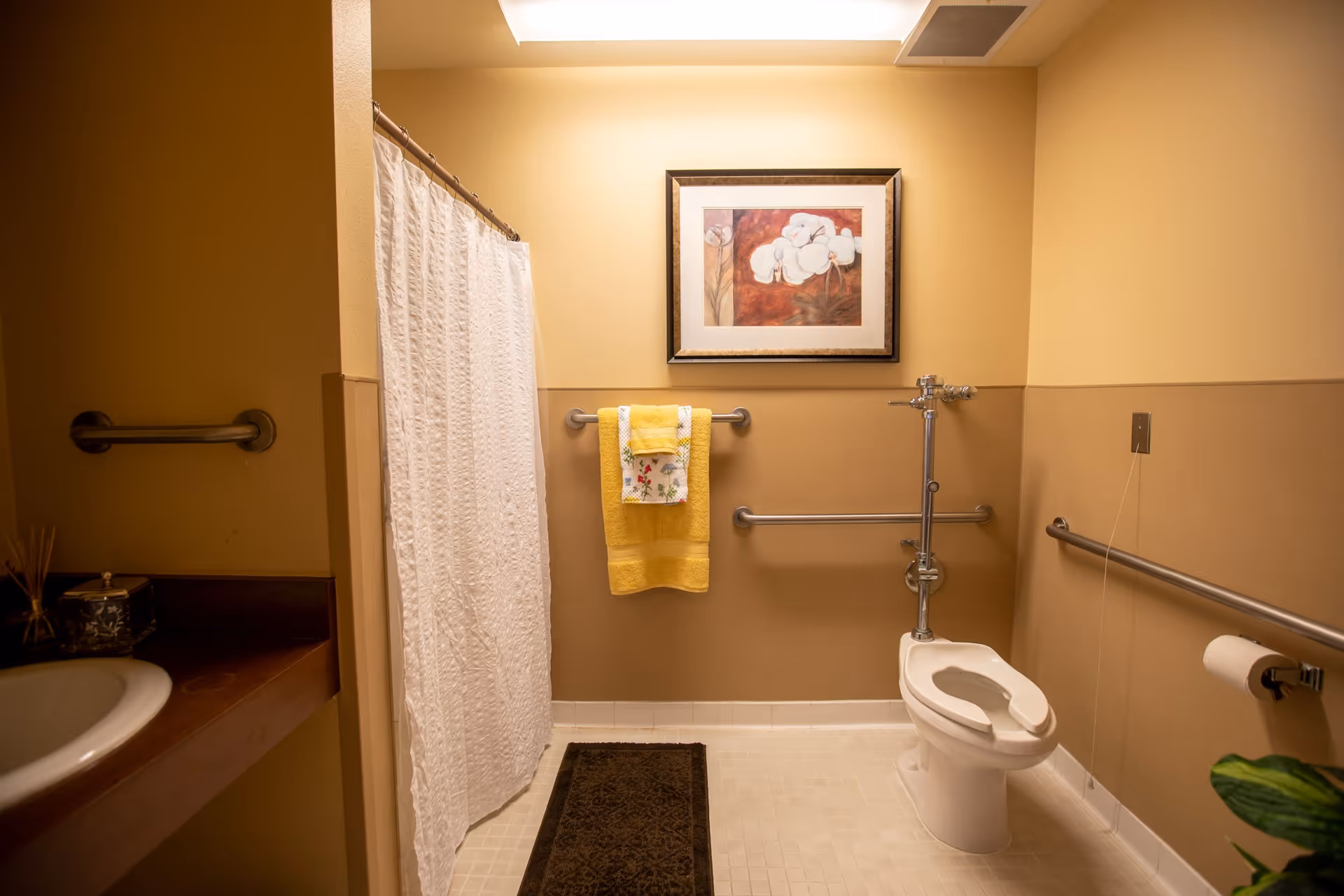 A senior living facility bathroom with beige walls and white tiled floor. The room features a toilet with support rails, a shower area with a white curtain, a sink with a countertop, and a towel rack holding yellow and floral towels. A framed floral artwork is hung above the towel rack.