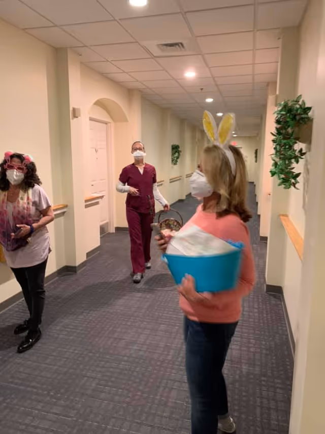Three women wearing face masks walking down a hallway in an assisted living facility. One woman in the foreground is wearing bunny ears and holding a blue basket filled with items. The hallway has beige walls, a gray carpet, and green plants mounted on the walls.