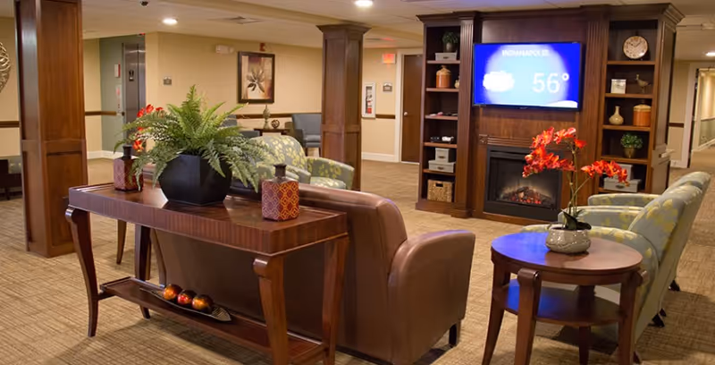 A cozy senior living community lounge area with comfortable armchairs and sofas arranged around a wooden coffee table and side table with a vase of red flowers. A wooden console table with decorative items and a large potted fern is behind a brown leather sofa. In the background, a built-in wooden entertainment center with a fireplace and a TV screen displaying the weather for Indianapolis at 56 degrees is visible. The room has warm lighting and beige carpeting.