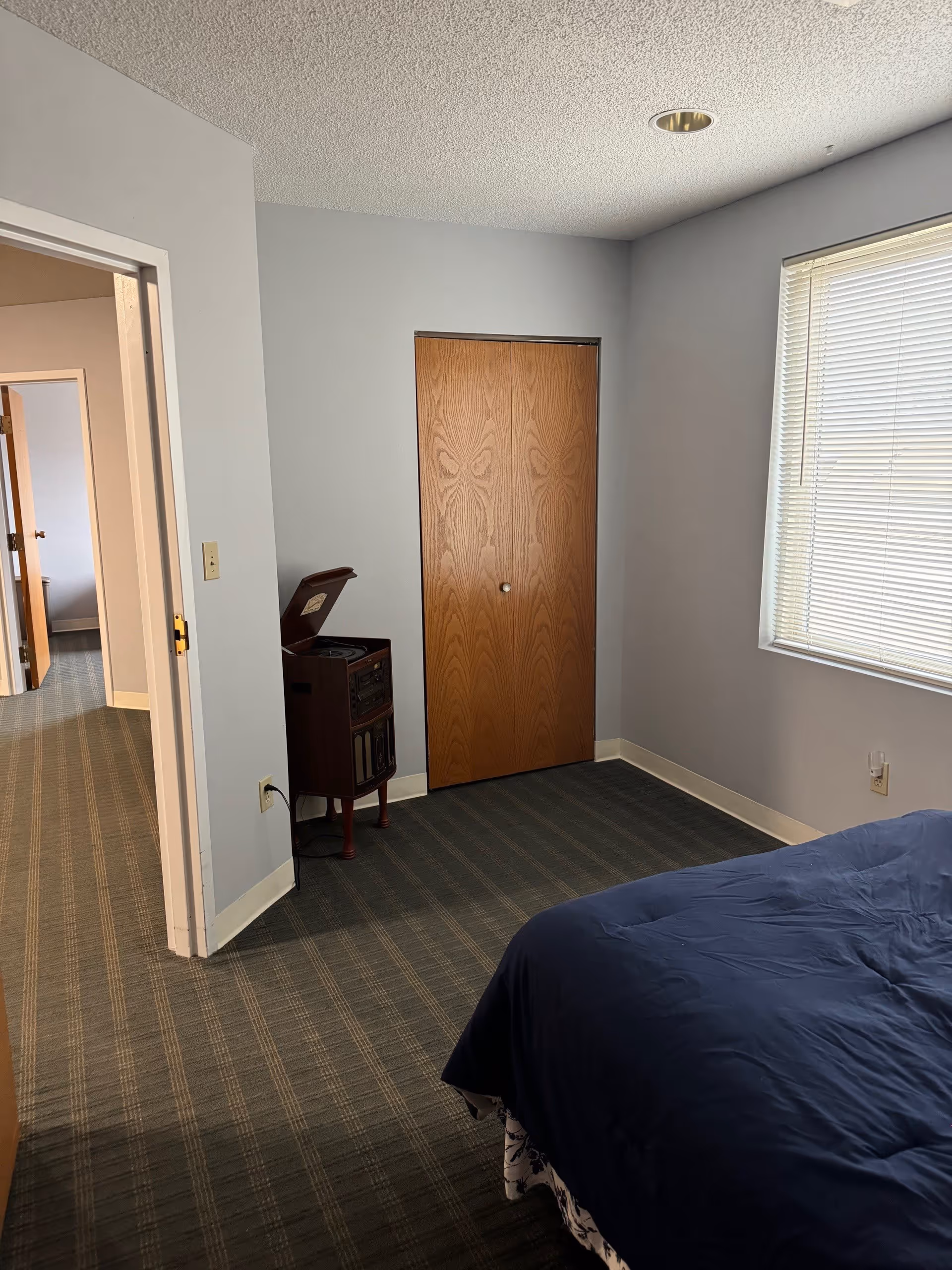 A bedroom with a blue bedspread partially visible on the right, a window with closed blinds on the right wall, a wooden closet door in the center, and an open doorway leading to another room on the left. There is a vintage-style record player or music system on a small stand near the closet. The room has light gray walls and a patterned carpet.