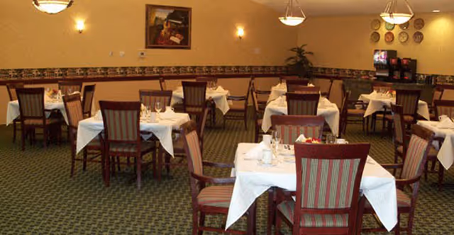 A dining room with multiple tables covered in white tablecloths, each set with glasses and napkins. The room has patterned carpet, beige walls with decorative trim, framed artwork, and wall-mounted light fixtures. There are several wooden chairs with striped upholstery around the tables.