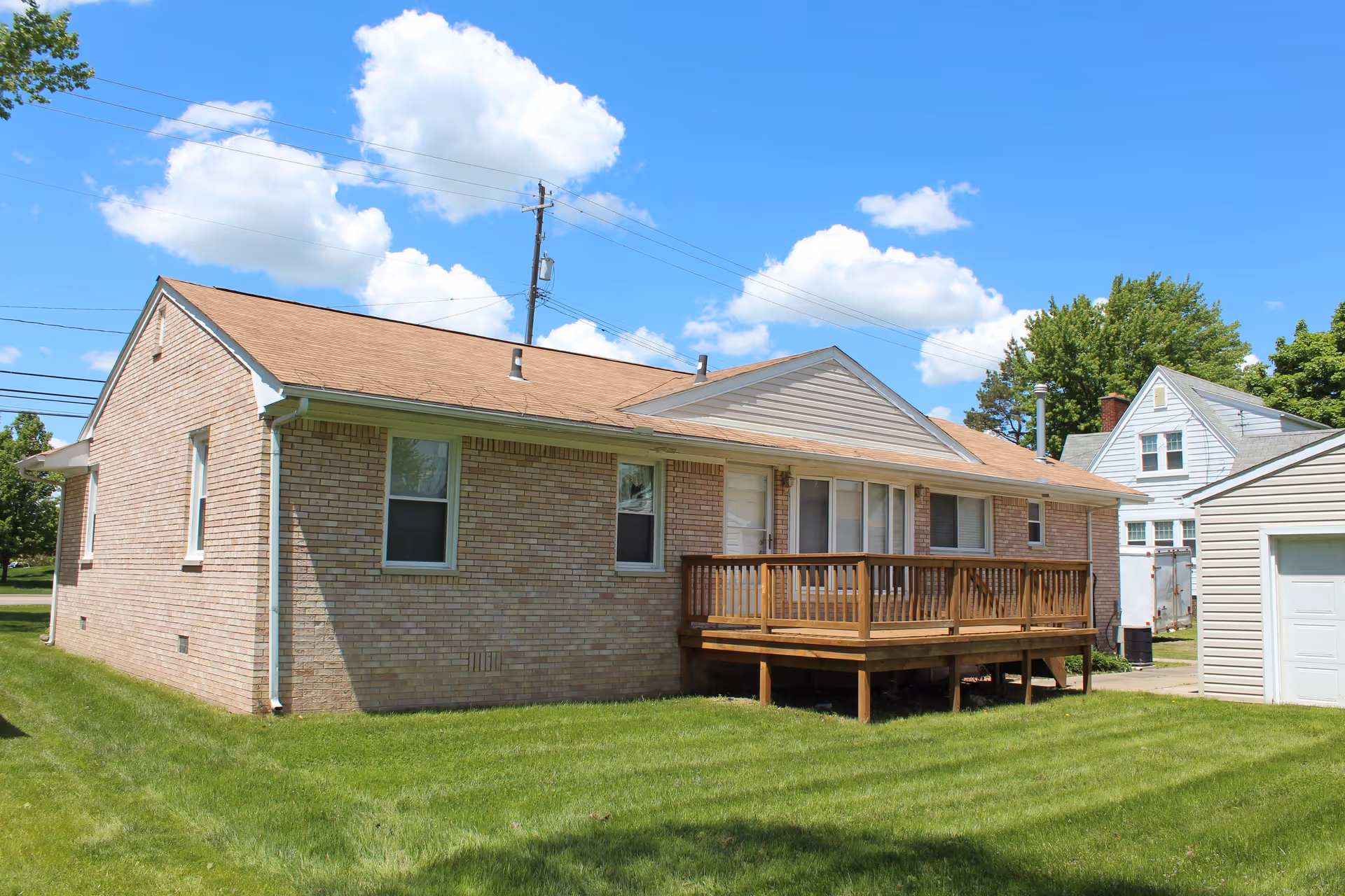 Exterior view of a single-story brick building with a small wooden deck and a grassy lawn under a blue sky with scattered clouds. There are power lines and neighboring houses visible in the background.