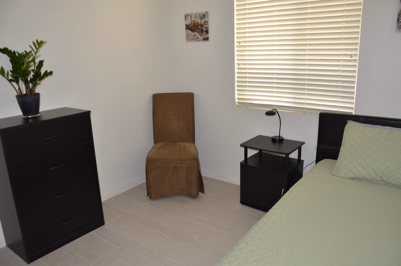 Small, minimally furnished bedroom with a bed, nightstand and lamp, dresser with a plant, and a chair by a window with closed blinds.