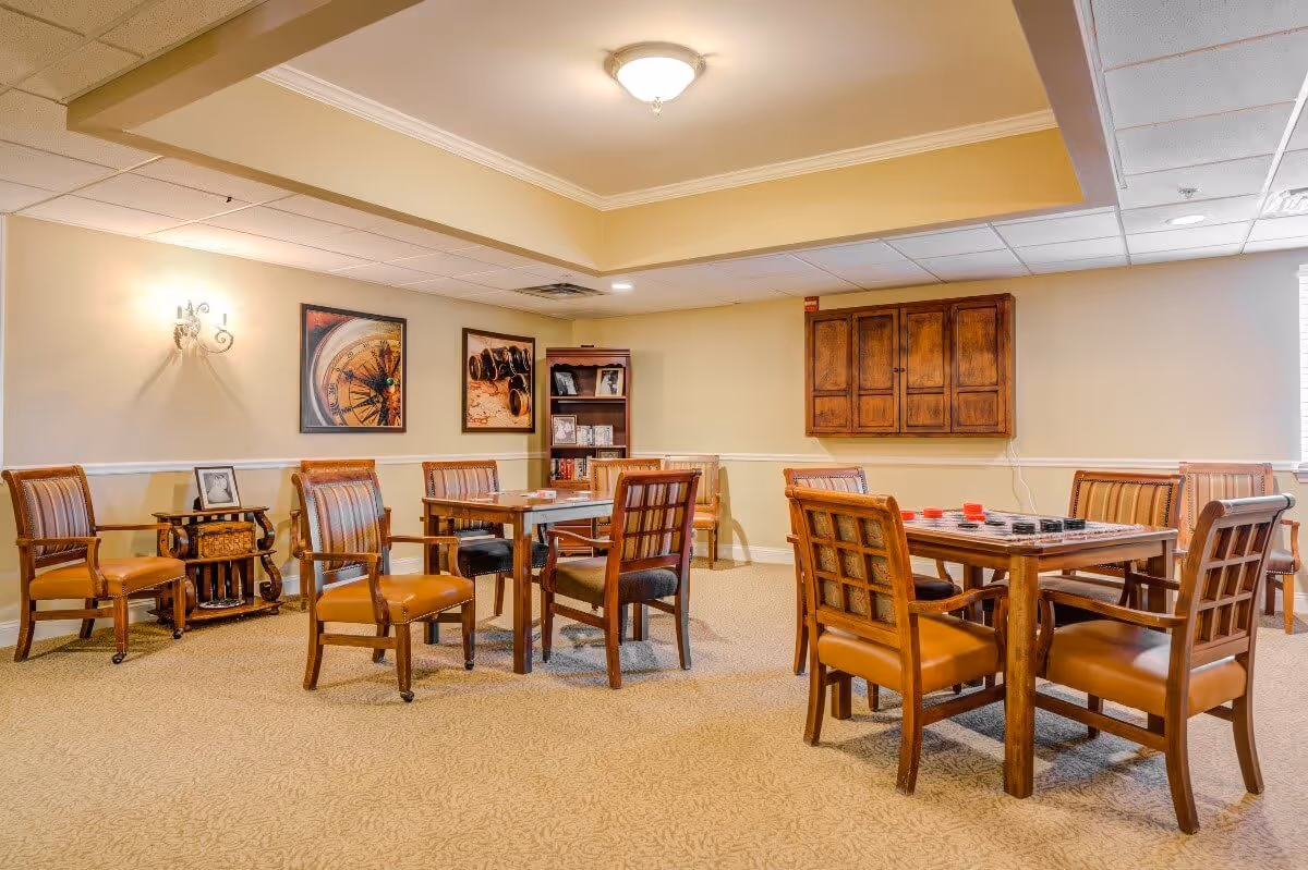 A well-lit room with beige walls and carpeted floor featuring multiple wooden tables and chairs arranged for group activities. The tables have game pieces on them, and the walls are decorated with framed pictures and a wooden cabinet. There is a small bookshelf in the corner with books and framed photos.