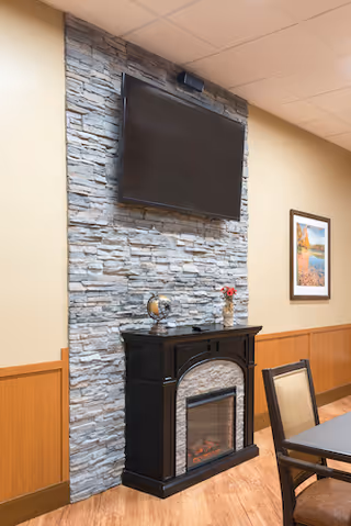 Wall-mounted TV above a stone-accented electric fireplace in a communal room with a framed picture and a table with chairs.