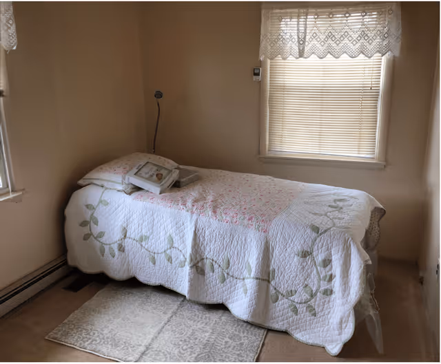 Small bedroom with a single bed covered by a white floral quilt, a picture frame on the pillow, and a window with blinds and lace curtains.
