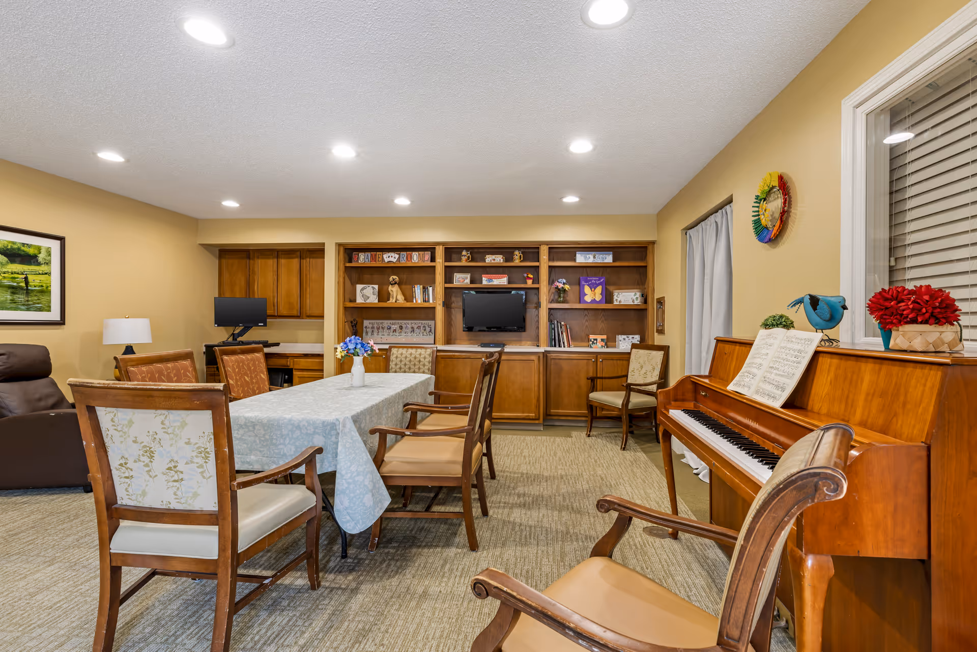 A cozy common room with a rectangular table covered with a light blue tablecloth surrounded by wooden chairs. To the right, there is a wooden piano with sheet music and decorative items on top. The back wall features built-in wooden shelves with books, decorations, and a small TV. The room has beige walls, carpeted floor, and recessed ceiling lights.