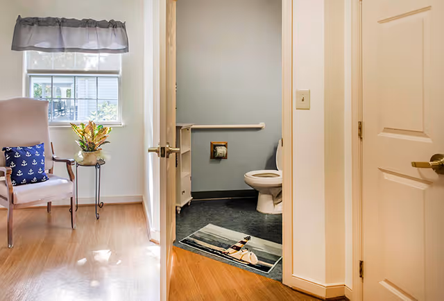 View of a bathroom with an open door showing a toilet, a small white cabinet, a grab bar on the wall, and a lighthouse-themed rug on the floor. Adjacent to the bathroom is a room with a wooden floor, a window with a gray valance, a beige armchair with a navy blue pillow featuring white anchors, and a small round table with a potted plant.