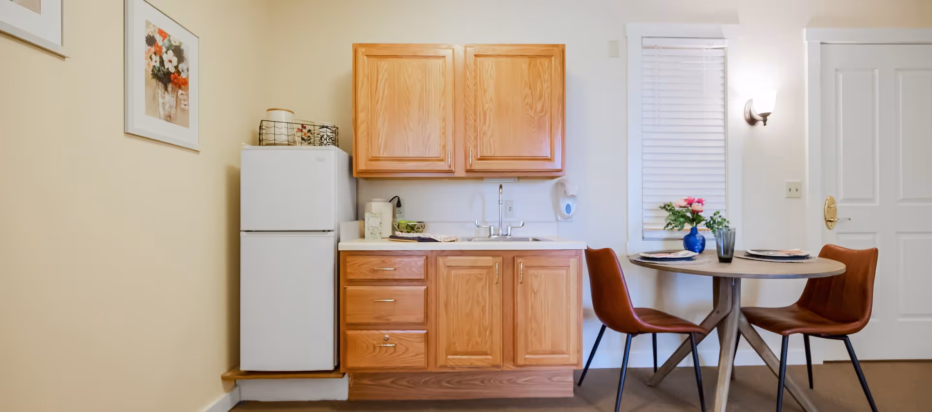 Small kitchenette with wooden cabinets, a refrigerator, sink, and a round dining table with two chairs.