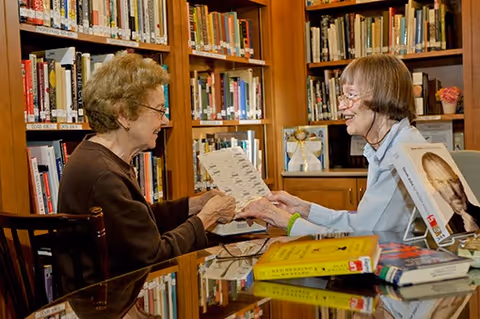Two elderly women sitting at a glass table in a library or reading room, smiling and holding a piece of paper. Bookshelves filled with books are visible in the background, along with several books on the table.