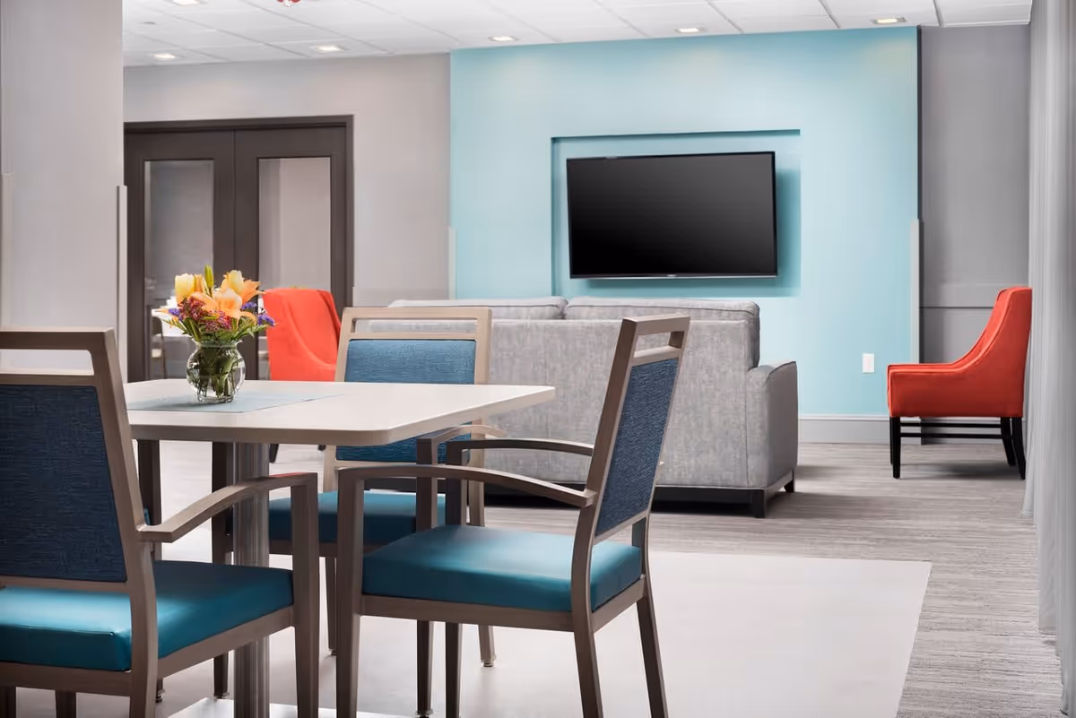 A modern and clean common area with a white table surrounded by four chairs with blue cushions. A small vase with colorful flowers is placed on the table. In the background, there is a gray sofa facing a wall-mounted flat-screen TV on a light blue accent wall. Two red armchairs are positioned near the sofa, and double doors with glass panels are visible on the left side.