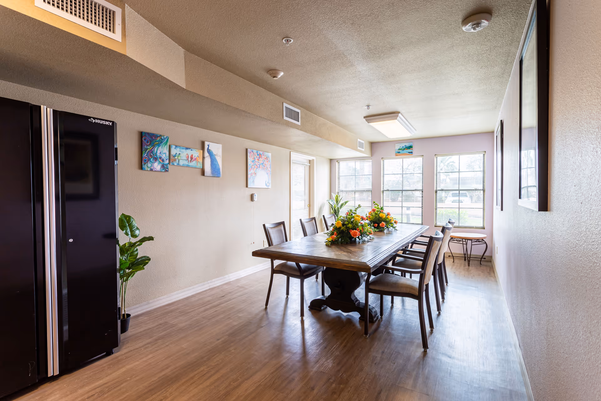 Bright dining room with a long wooden table, chairs, a floral centerpiece, and windows at the far end.