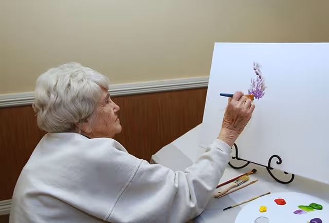 An elderly woman with white hair is painting on a white canvas set on a stand. She is using a paintbrush to create purple and pink floral shapes. On the table in front of her are several paintbrushes and a palette with various colors of paint.