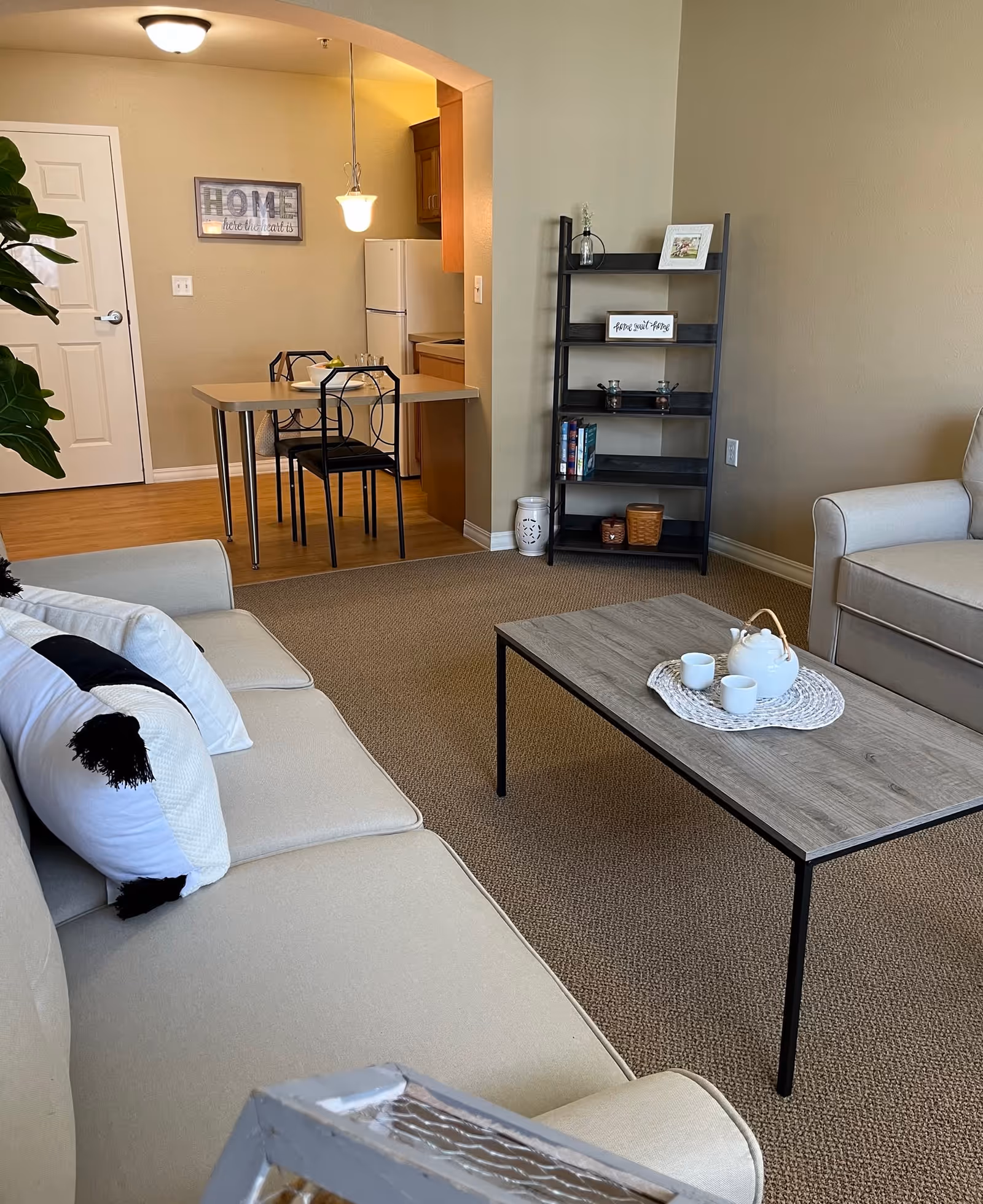 A cozy living room and dining area in a senior living facility. The living room features a beige sofa with white and black pillows, a wooden coffee table with a white teapot and two cups on a woven placemat, and a black ladder-style shelf with decorative items and books. The dining area has a small table with two black metal chairs, a white refrigerator, wooden cabinets, and a wall sign that reads 'HOME is where the heart is'. The walls are painted beige and the floor is carpeted in the living room and wood in the dining area.
