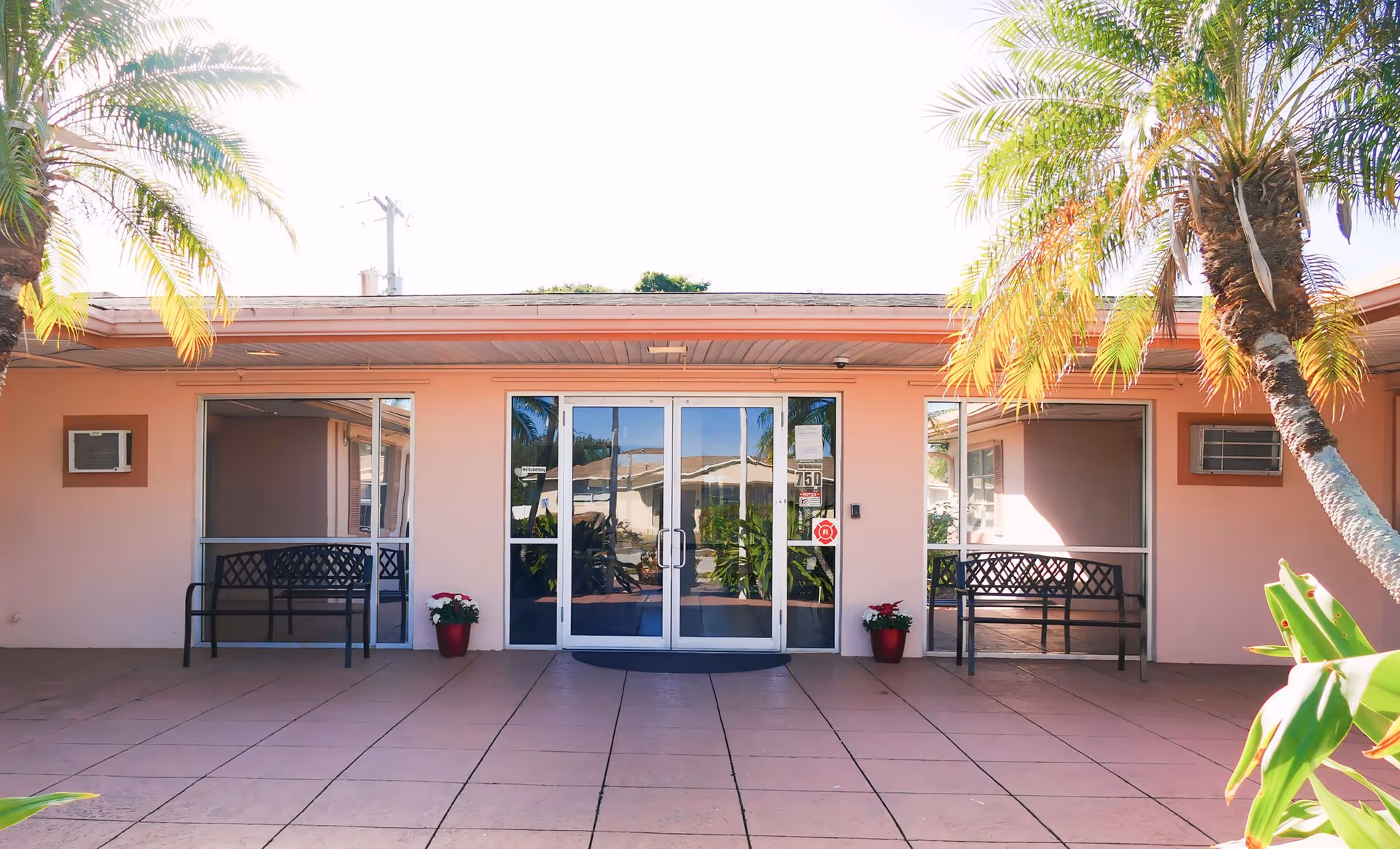 Glass double-door entrance to a single-story pink care center building flanked by benches, potted plants, and palm trees.