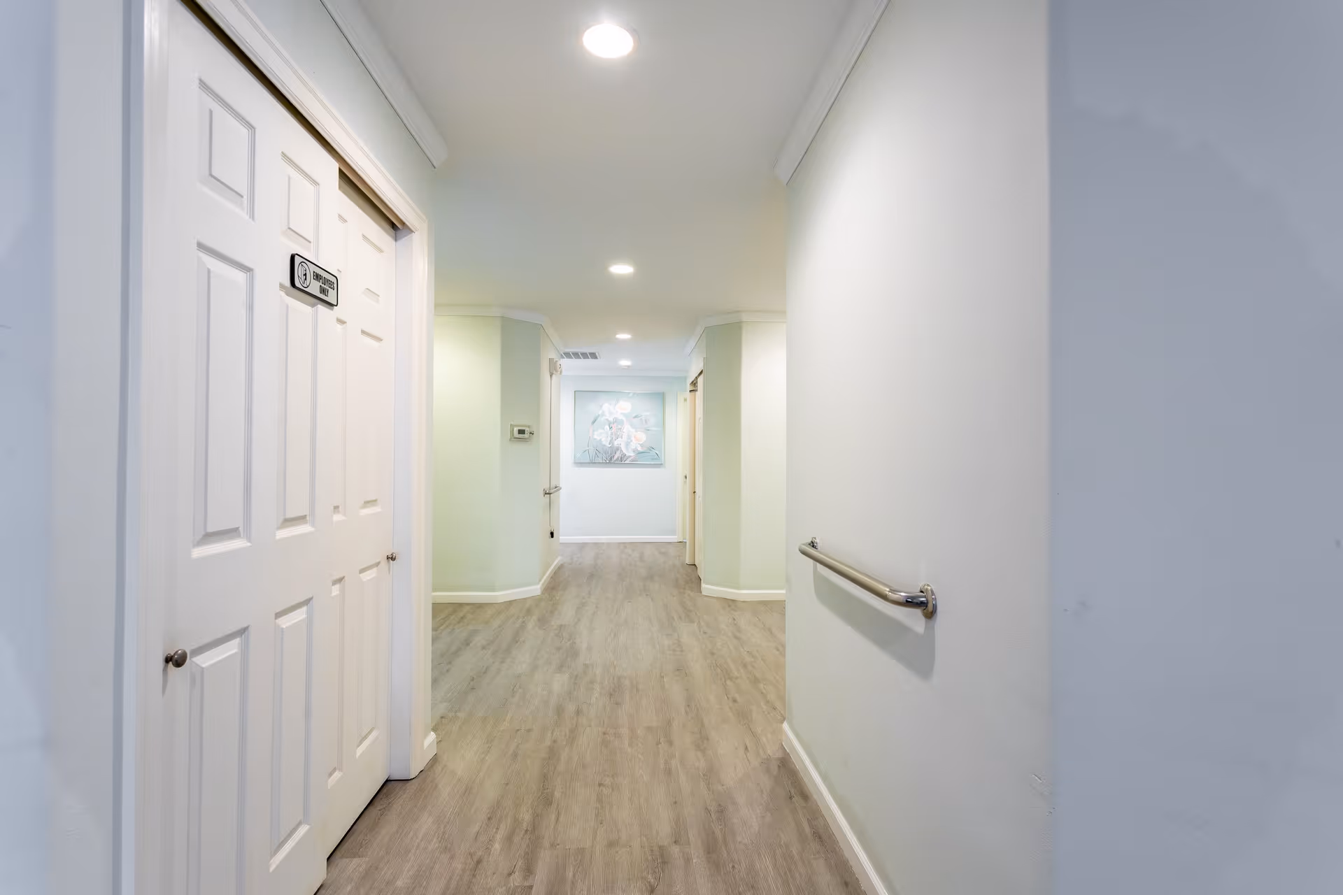A clean, well-lit hallway with light-colored walls and wood flooring. There is a handrail on the right wall and a door on the left labeled 'Employees Only'. At the end of the hallway, a floral painting is visible on the wall.
