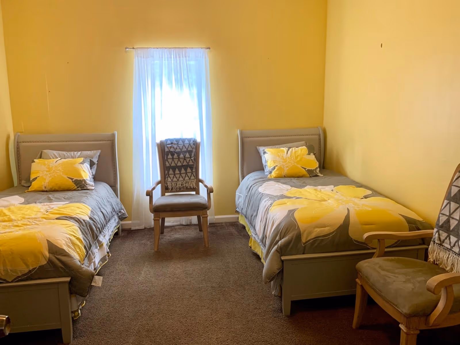 A bedroom with two single beds placed against opposite walls, each bed has gray and yellow floral bedding and matching pillows. Between the beds is a window with sheer white curtains, and in front of the window is a wooden chair with a patterned throw draped over the back. Another wooden chair with a similar patterned throw is partially visible on the right side of the image. The walls are painted yellow and the floor is carpeted in brown.