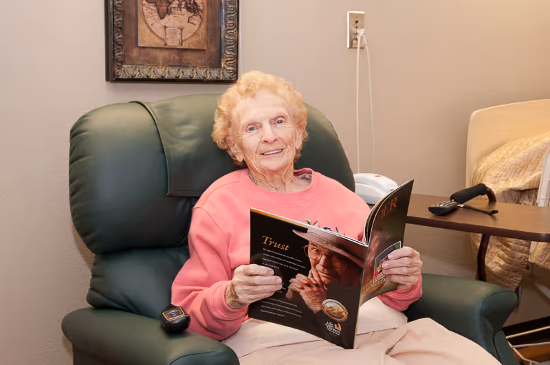 An elderly woman with curly blonde hair wearing a pink sweatshirt is sitting in a green recliner chair, smiling and holding an open magazine. Behind her is a beige wall with a framed picture and an electrical outlet. To the right, there is a hospital-style bed with a beige blanket and a wooden overbed table holding a TV remote.