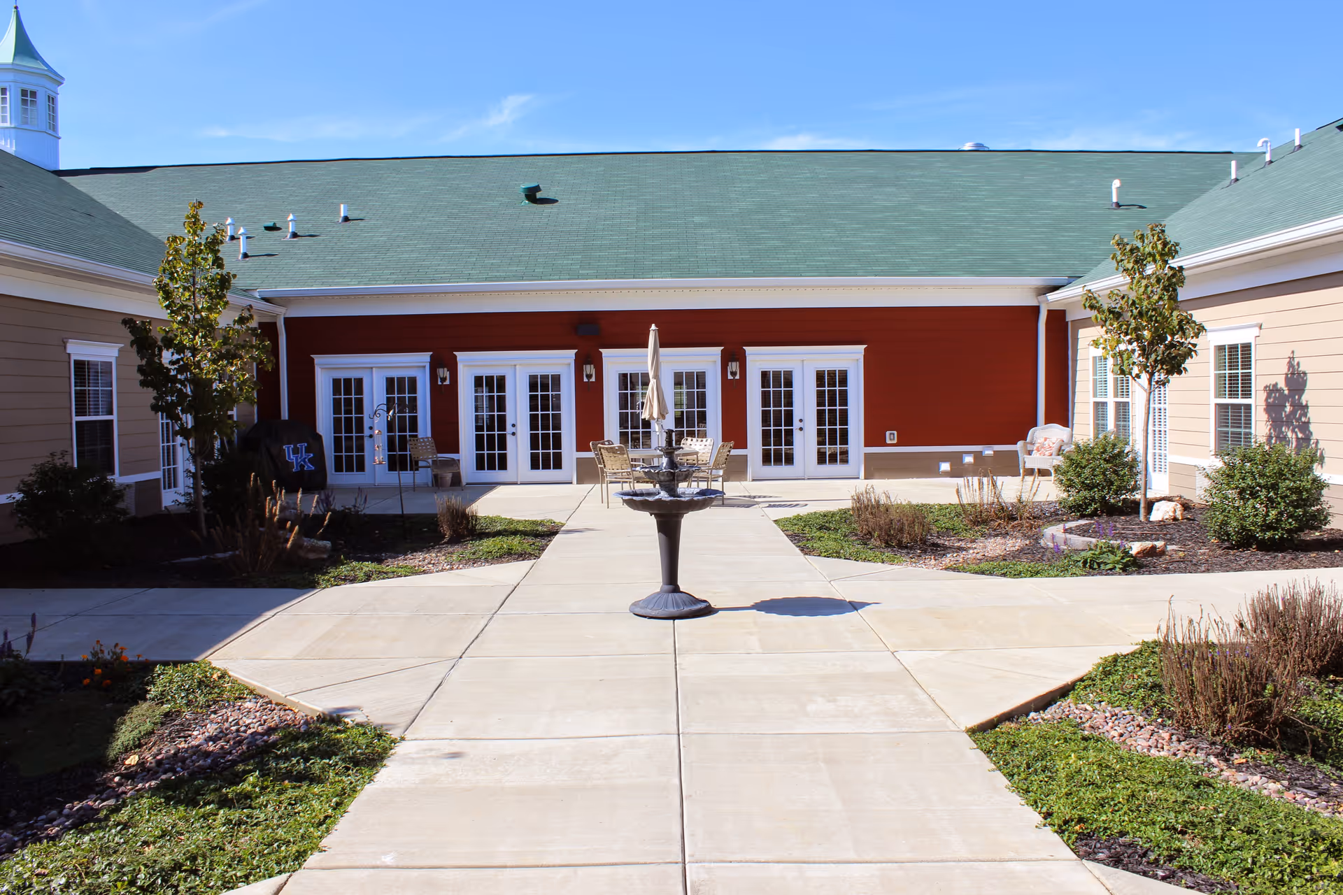 Outdoor courtyard area of a senior living facility with a central fountain, surrounded by paved walkways, small landscaped garden beds, and buildings with multiple glass doors and windows under a green roof.