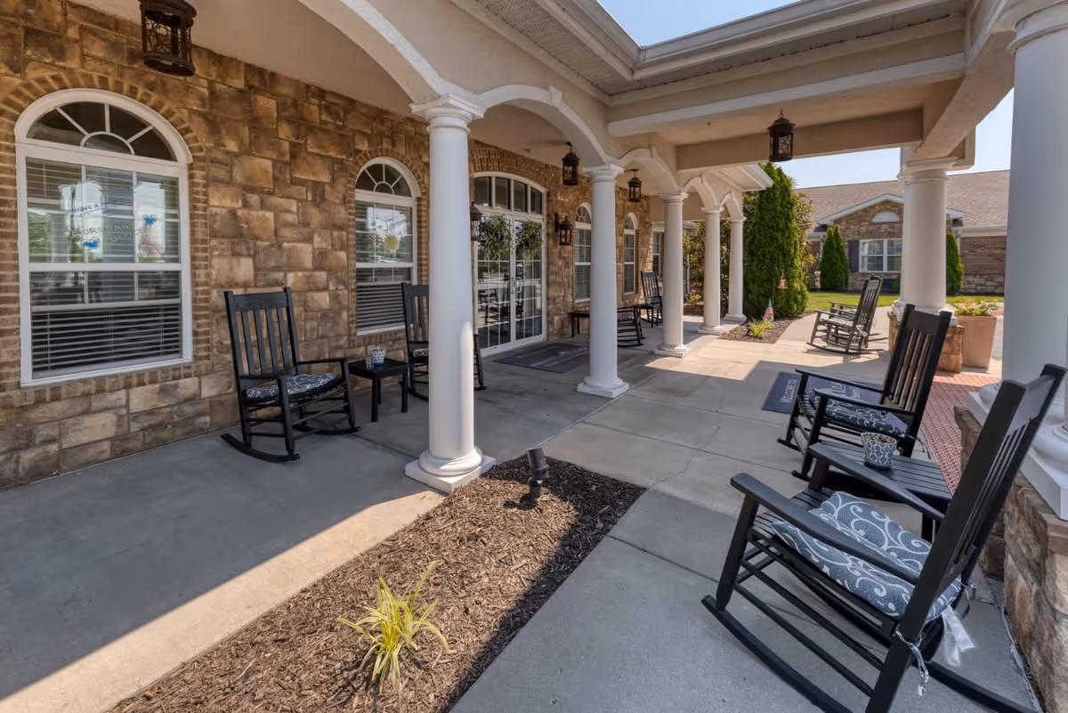Covered outdoor patio area with stone walls and white columns, featuring several black rocking chairs with cushions and small tables. The patio overlooks a landscaped garden area with shrubs and trees, under a clear blue sky.