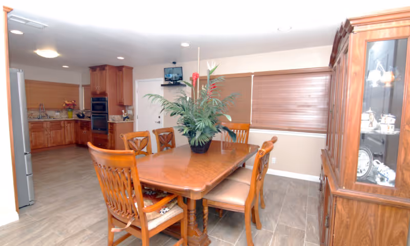 A dining room with a wooden dining table and six matching chairs. A large potted plant is centered on the table. To the right is a wooden china cabinet with glass doors displaying dishes. In the background, there is a kitchen area with wooden cabinets, a refrigerator, and an oven. The room has tiled flooring and windows with closed blinds.
