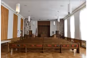 Interior view of a chapel or worship room with wooden pews arranged in rows facing a wooden altar at the front. The room has large windows on the right side allowing natural light to enter, pendant lights hanging from the ceiling, and wooden paneling on the walls.