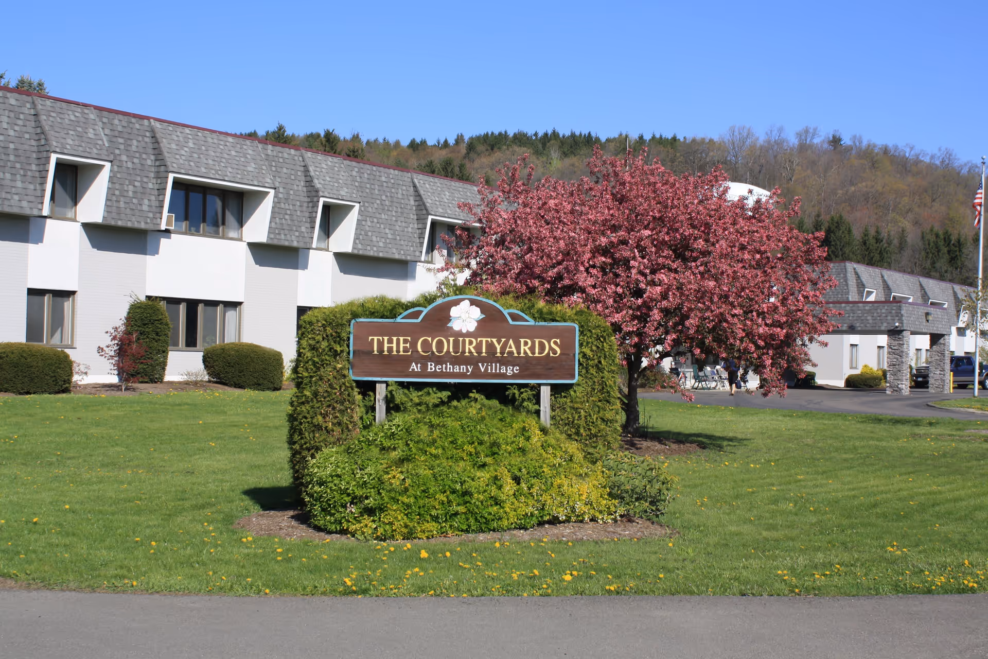 A wooden sign reading "The Courtyards At Bethany Village" sits on a landscaped lawn in front of a two-story building and a pink-flowering tree.