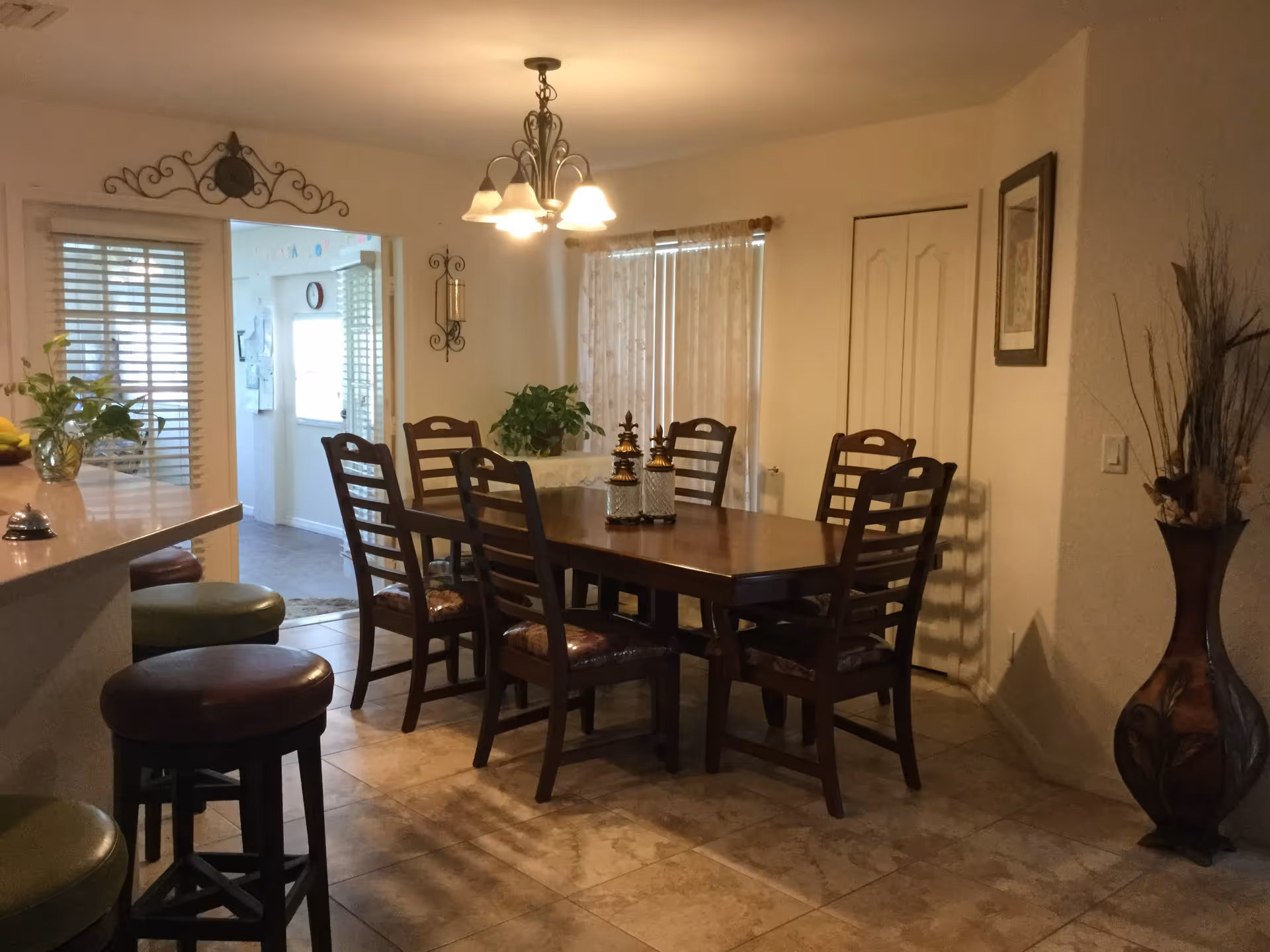 Dining room with a wooden table and six chairs under a chandelier, bar stools at a counter, and decorative plants and vases.