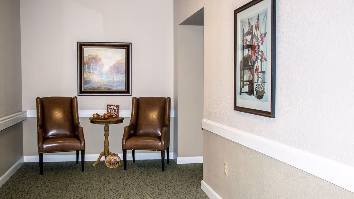 A small seating area in a hallway with two brown leather chairs and a round wooden table between them. On the table is a decorative sign and some fall-themed decor. There is a framed landscape painting on the wall behind the chairs and another framed artwork on the adjacent wall. The floor is carpeted with a green patterned carpet.