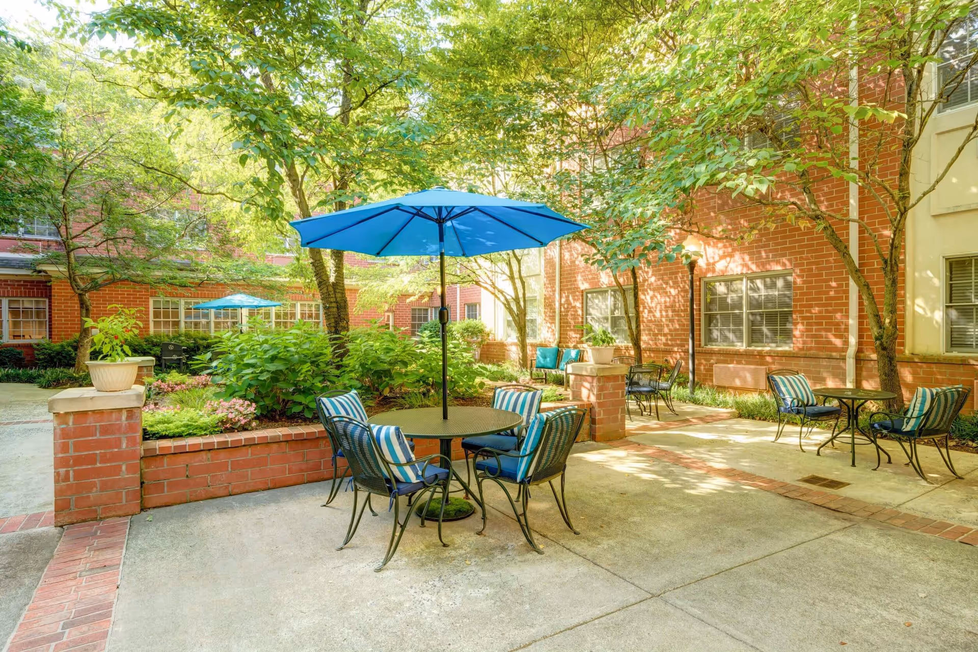 Outdoor courtyard area at Magnolia Place of Roswell with several round tables and chairs, some with blue umbrellas. The space is surrounded by brick buildings and lush green trees and plants, creating a shaded and inviting atmosphere.