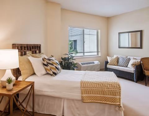 A bright and cozy bedroom with a neatly made bed featuring white linens and patterned pillows. A wooden nightstand with a lamp and small plant is beside the bed. There is a window with blinds, a small air conditioning unit below it, and a wicker loveseat with cushions against the wall. A framed mirror hangs above the loveseat.