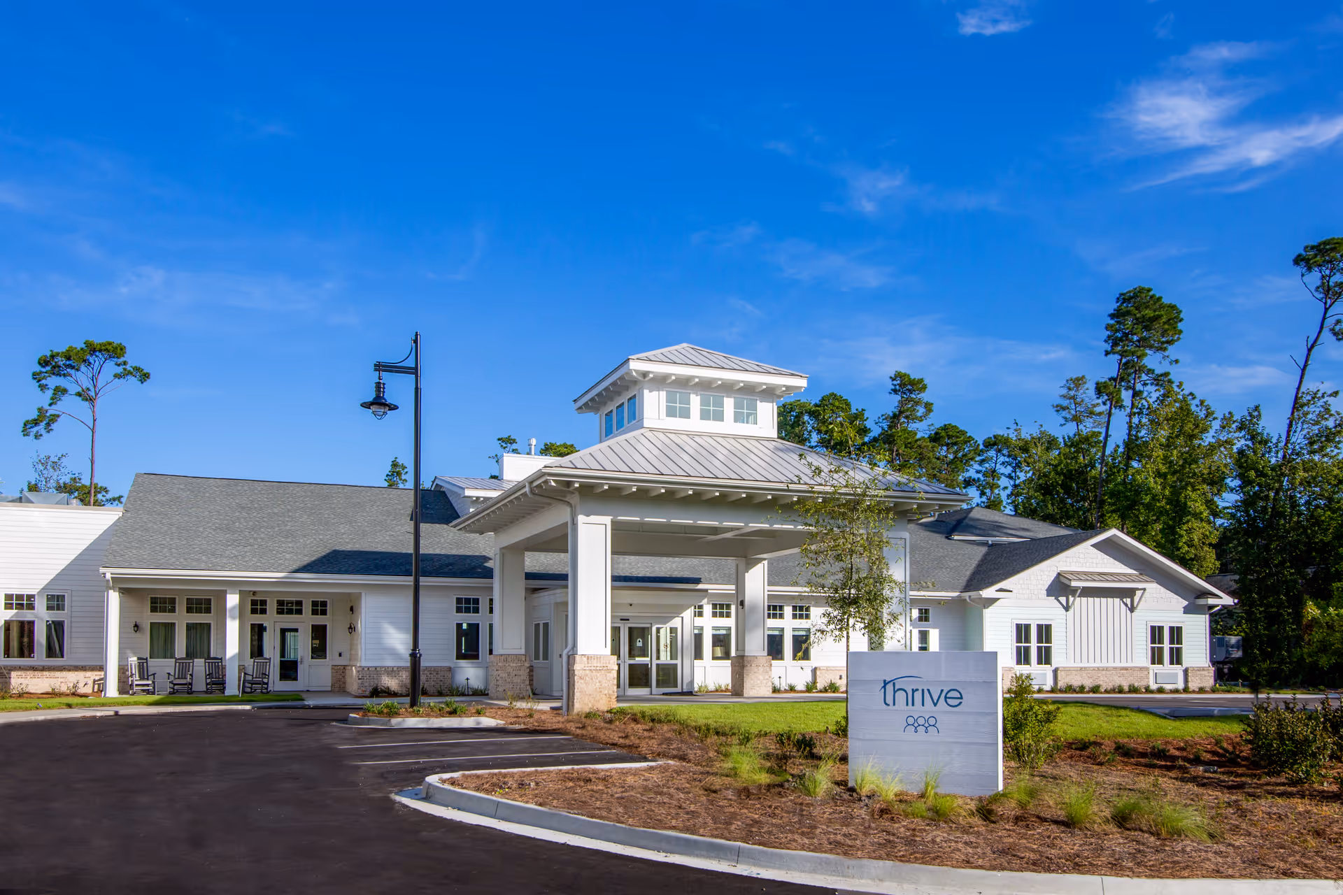 Exterior view of a single-story white building with a covered entrance and a sign in front that reads 'Thrive'. The building is surrounded by a paved driveway, landscaped grass, and trees under a clear blue sky.