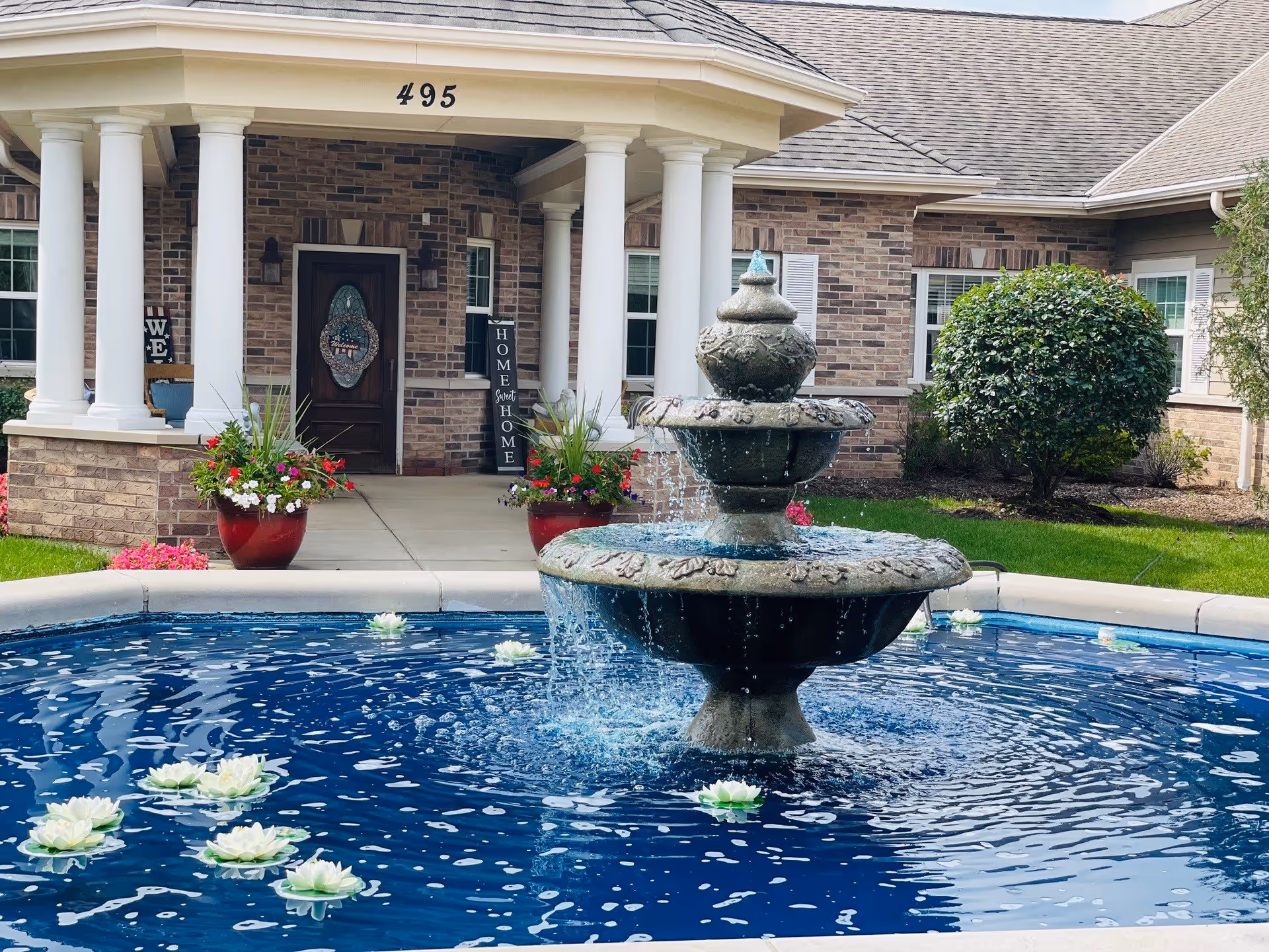 Outdoor view of the entrance to a brick building with white columns and the number 495 above the door. In front of the entrance is a large stone fountain with water flowing and white floating flowers in the water. There are potted plants with flowers on either side of the walkway leading to the door, and green bushes and grass surround the area.
