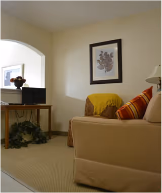 Small living area with a beige sofa and striped pillows, framed artwork on the wall, a side lamp, and a desk with a small TV and plant.