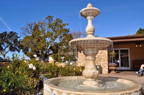 Outdoor scene at Huntington Valley Healthcare featuring a large stone water fountain with water flowing from the top tier to the basin below, surrounded by greenery and white flowers. In the background, there is a building with a covered patio area and glass doors, under a clear blue sky.