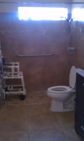 Bathroom with a toilet, a grab bar on the wall, a showerhead, and a shower chair. The walls are tiled and there is a window near the ceiling letting in light.