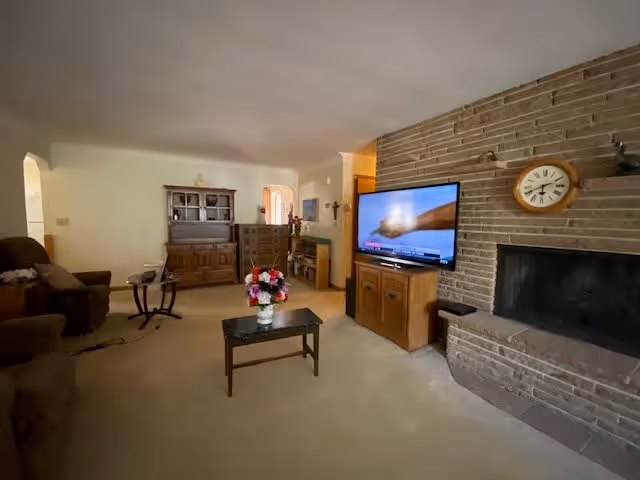 A cozy living room with beige carpeting, a brick fireplace with a clock above it, a flat-screen TV on a wooden cabinet, a coffee table with a flower arrangement, and several pieces of wooden furniture including a hutch and a small table. There are armchairs on the left side and an archway leading to another room.