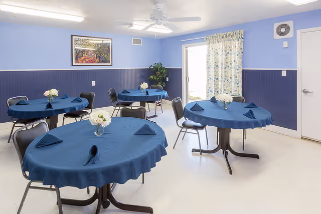 A small dining room with four round tables covered with blue tablecloths and matching blue napkins folded on each table. Each table has a small vase with white flowers. The room has light blue walls with a darker blue wainscoting, a ceiling fan, a framed picture on the wall, a potted plant in the corner, and a door with floral curtains leading outside.