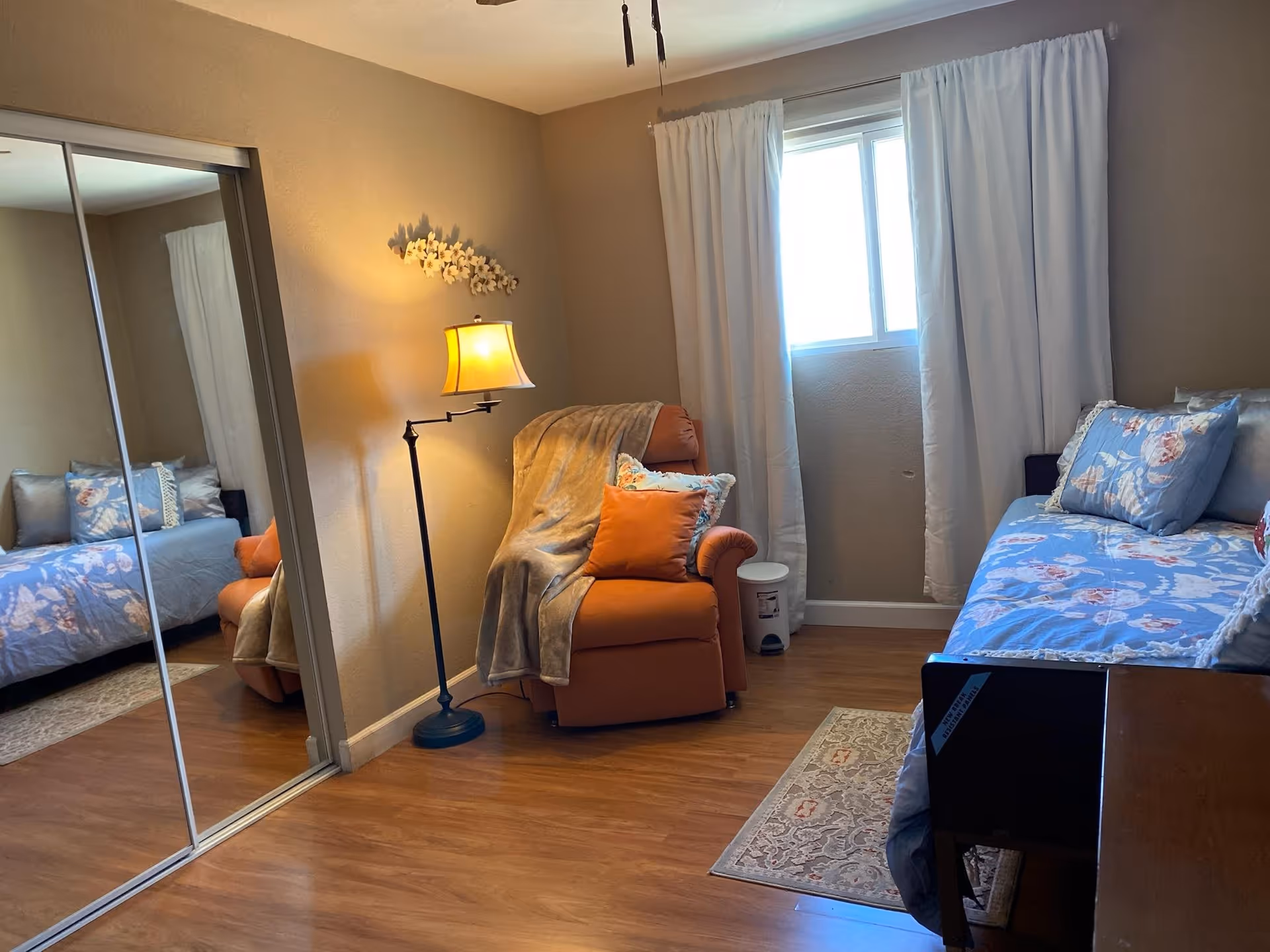 Cozy bedroom with an orange armchair and floor lamp, a daybed with floral bedding, mirrored closet doors, and a window with white curtains.