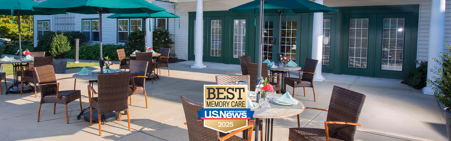 Outdoor patio area at Crescent Point at Niantic with several round tables and wicker chairs arranged for seating. Each table is set with napkins and small flower arrangements. Large green umbrellas provide shade over the tables. The building exterior features green doors and white columns.