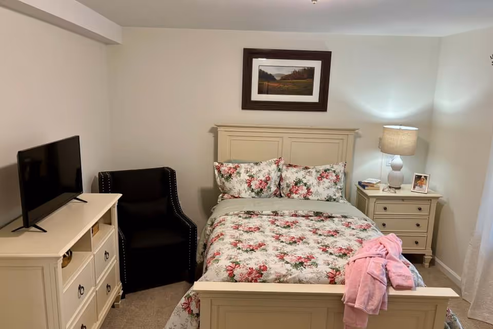 A cozy bedroom with a cream-colored wooden bed frame and floral bedding. A pink robe is draped over the footboard. To the right of the bed is a matching nightstand with a lamp, books, and a framed photo. To the left of the bed is a black armchair next to a cream-colored dresser with a flat-screen TV on top. A framed landscape picture hangs on the wall above the bed.