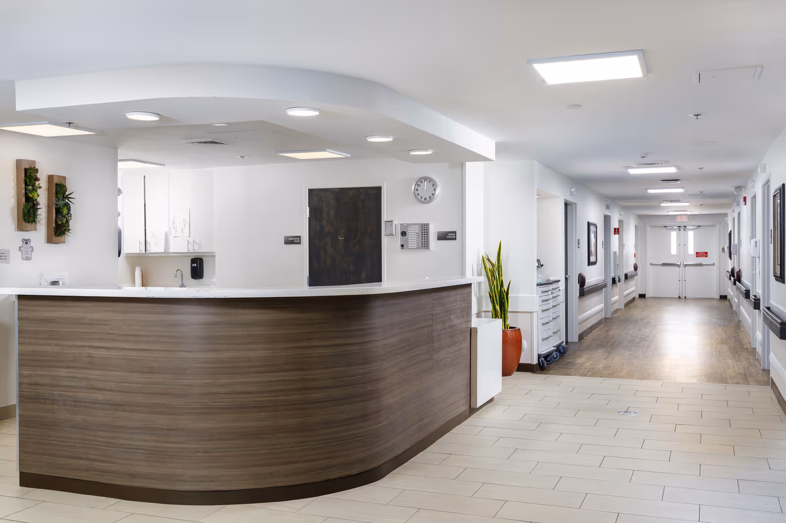 Reception desk area in a senior living facility with a curved wooden counter, white walls, ceiling lights, a clock, and a long hallway with multiple doors and framed pictures on the walls.